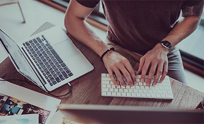 Person typing on a white keyboard at a wooden desk with a laptop nearby.