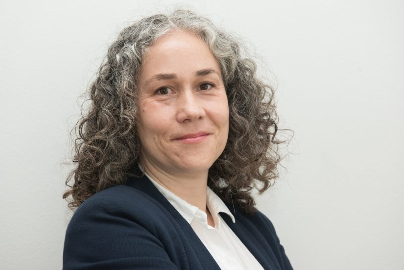 Professional headshot of a smiling woman with shoulder-length curly gray hair, wearing a navy blazer and white blouse against a light background.